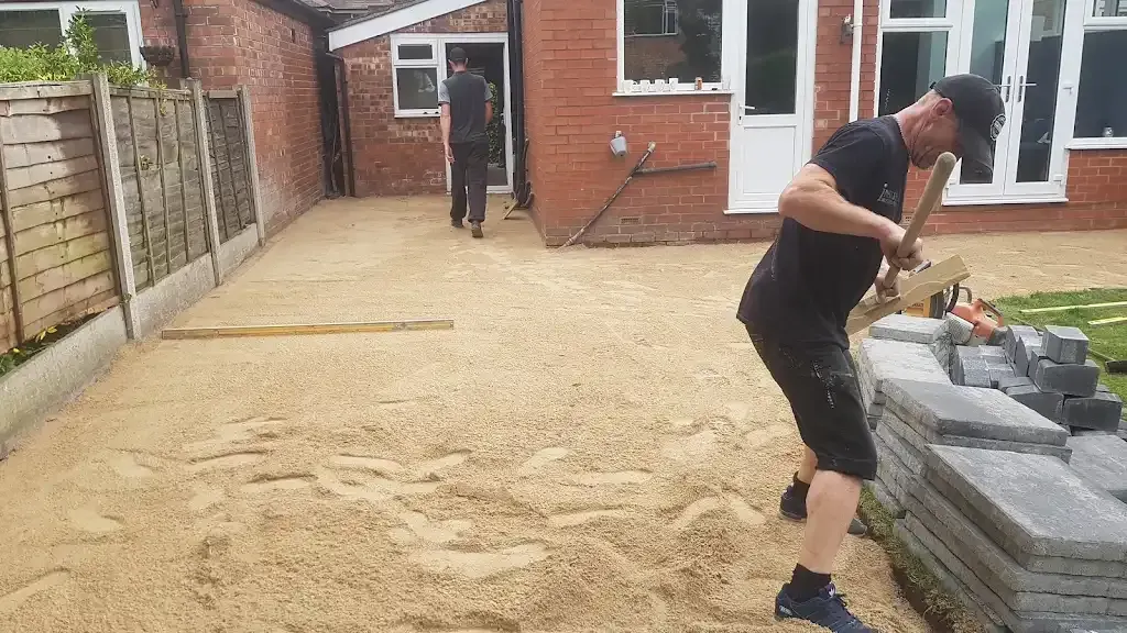 Workers installing a new block paved driveway or patio, preparing the base layer with sand and laying paving stones.